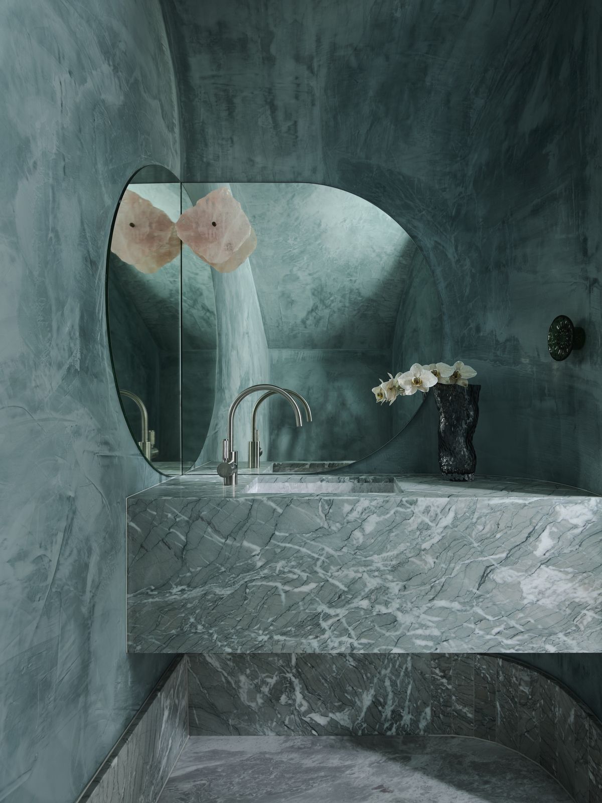 moody bathroom with emerald green venetian plaster walls, a green marble floating vanity, a corner mirror and chrome tapware, with a crystal wall sconce and vase filled with flowers