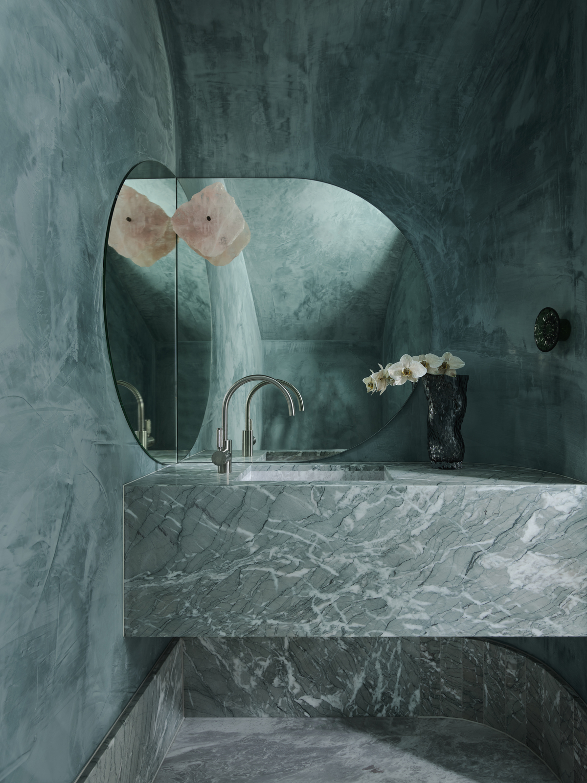 moody bathroom with emerald green venetian plaster walls, a green marble floating vanity, a corner mirror and chrome tapware, with a crystal wall sconce and vase filled with flowers