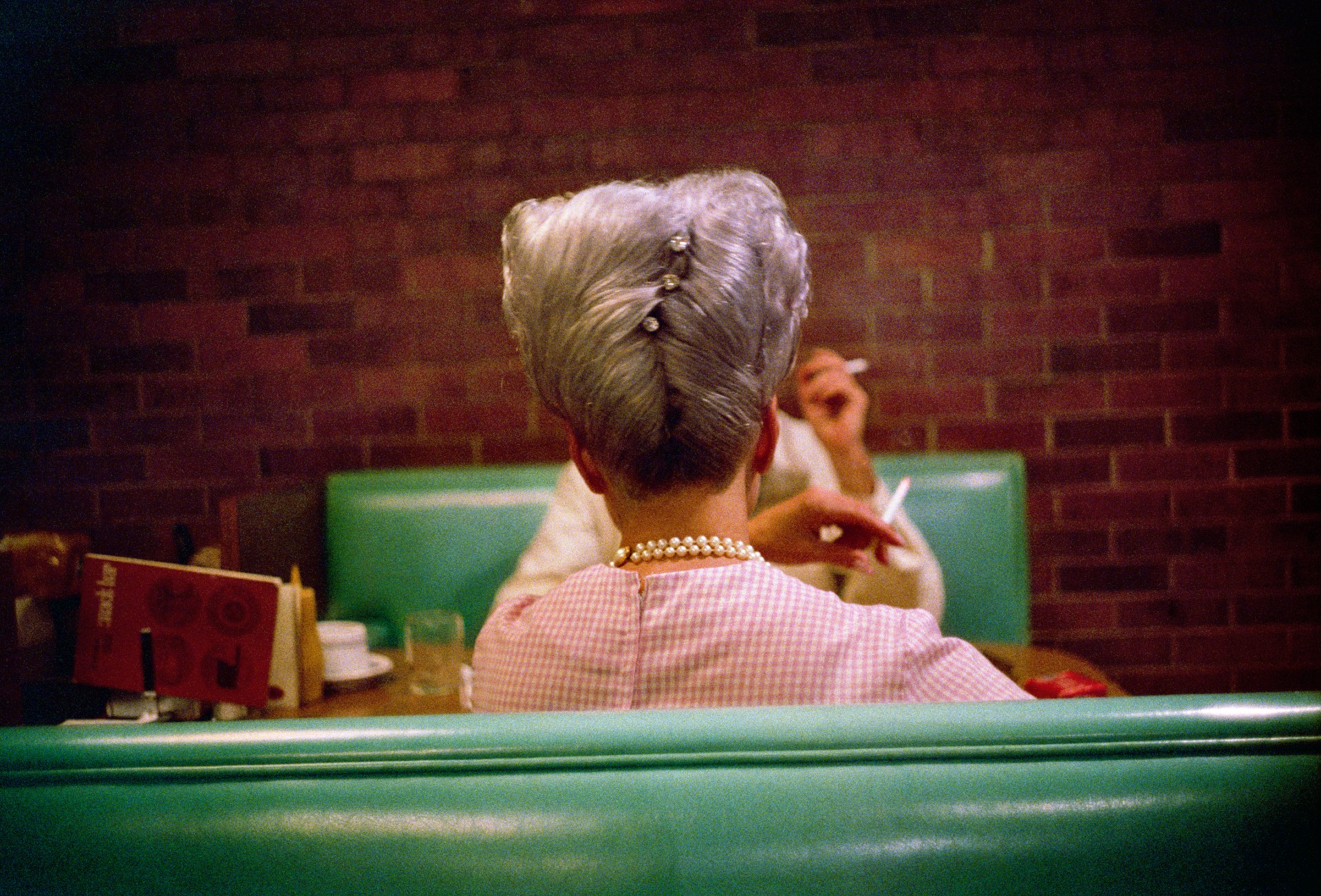A color photograph taken from behind a woman with grey hair swept into an updo, sitting in a diner booth as a man across the table smokes a cigarette.