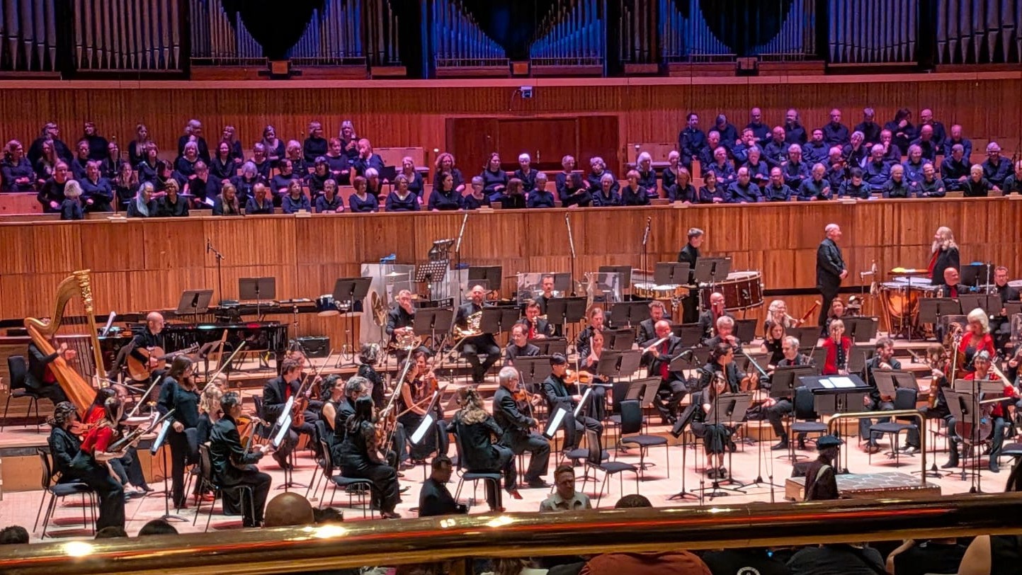 Stage full of musicians with string instruments, in the background there is a choir.