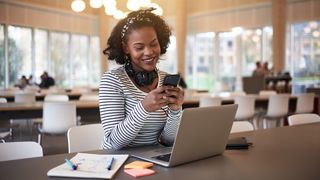 Student with headphones around her neck using a phone while sitting in a cafe in front of a laptop