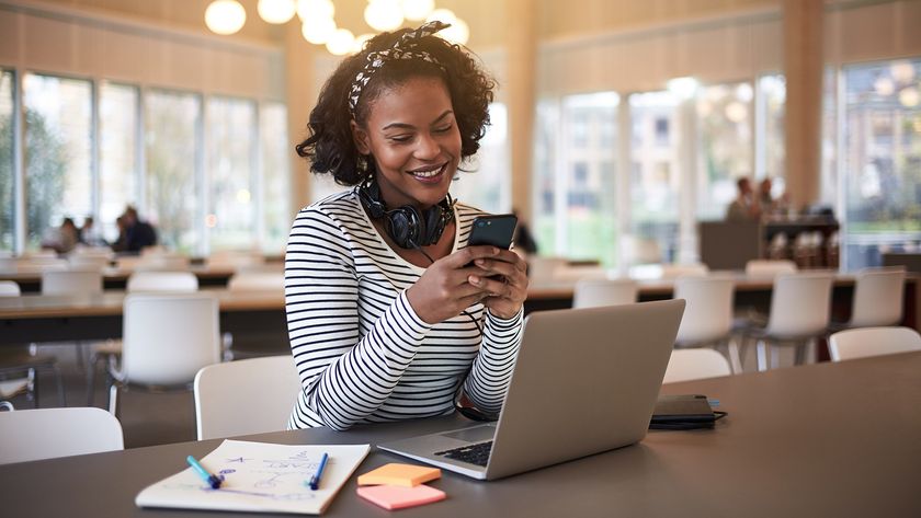 Student with headphones around her neck using a phone while sitting in a cafe in front of a laptop