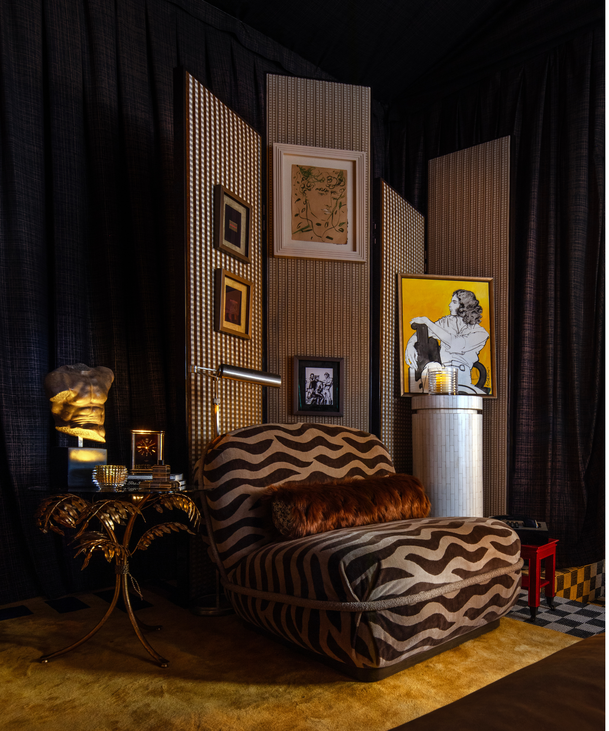 Moody sitting room vignette featuring a patterned folding screen dotted with tiny art, a brass palm tree-motif cocktail table, and an armless tiger-print slipper chair