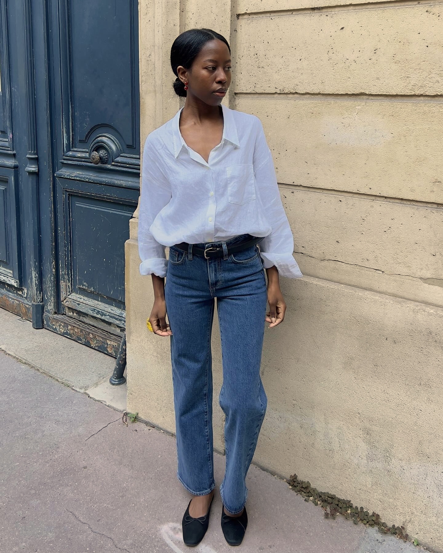 French style influencer poses on a Paris sidewalk wearing a white button-down shirt, black belt, straight-leg jeans, and black ballet flats