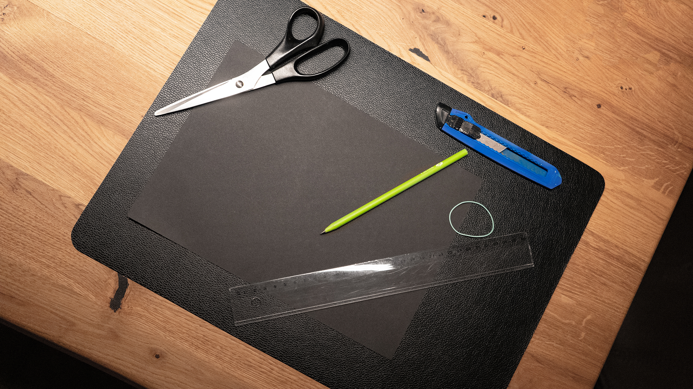 A workspace with black paper, scissors, a green pencil, a ruler, a blue utility knife, and an elastic band on a textured mat