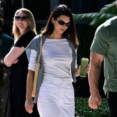Kendall Jenner walks outside wearing white straight-leg trousers with a white boat-neck t-shirt and black slipper shoes.