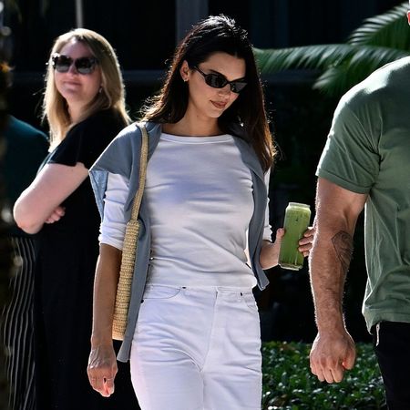 Kendall Jenner walks outside wearing white straight-leg trousers with a white boat-neck t-shirt and black slipper shoes.