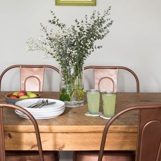 wooden dining table with copper industrial chairs and glass vase of green foliage