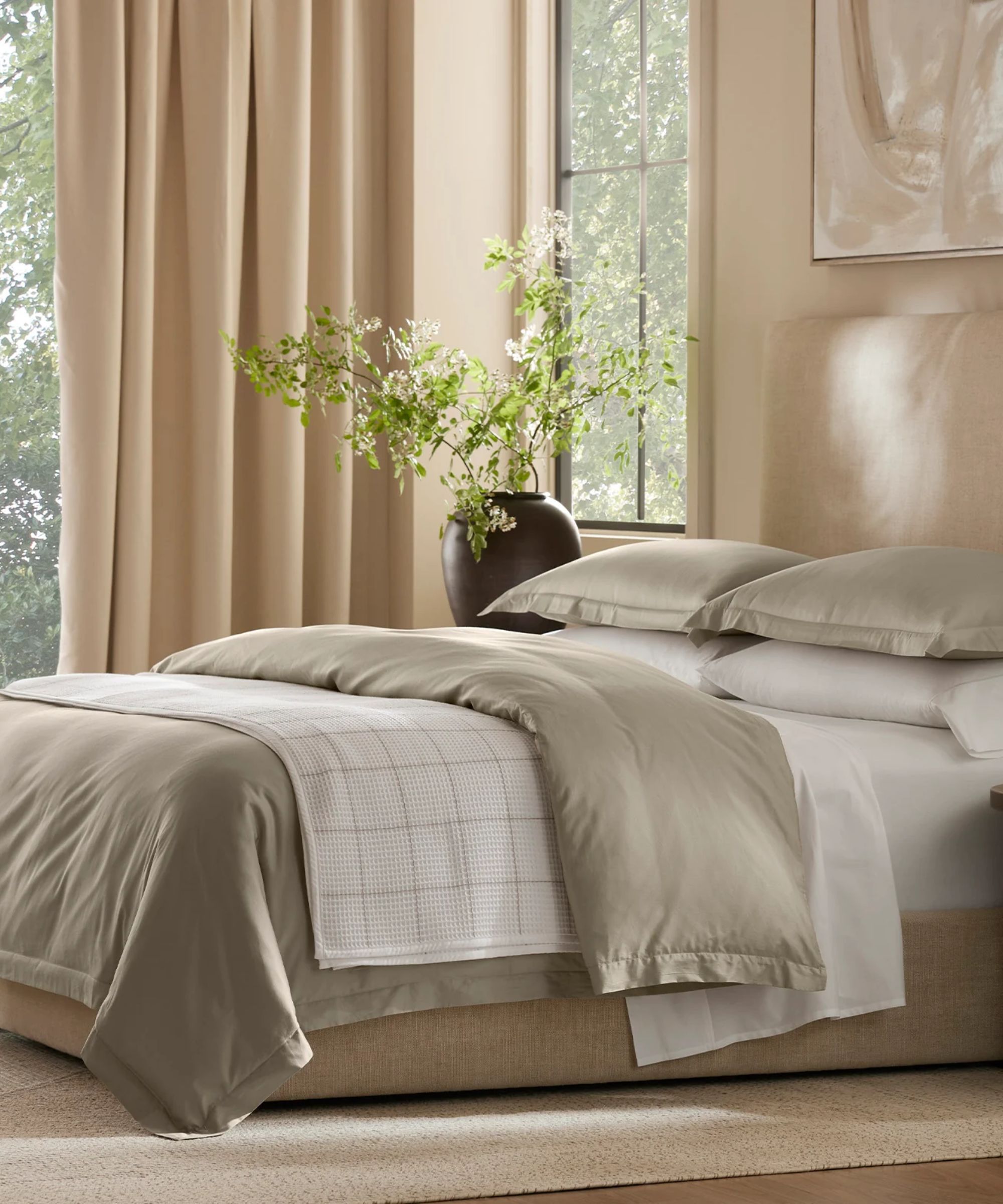 Neutral modern bedroom with cream bed, white and gray bedding, cream curtains, vase with plant stems