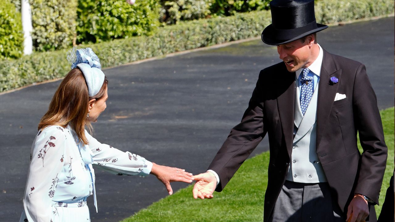 Carole Middleton wearing a pale blue floral dress and matching headband with a flower reaching her hand out to grab Prince William&#039;s while standing in grass at Royal Ascot 