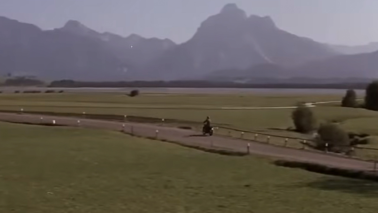 An incredible vista of fields and mountains with a road running trough it and a man on motorcycle.