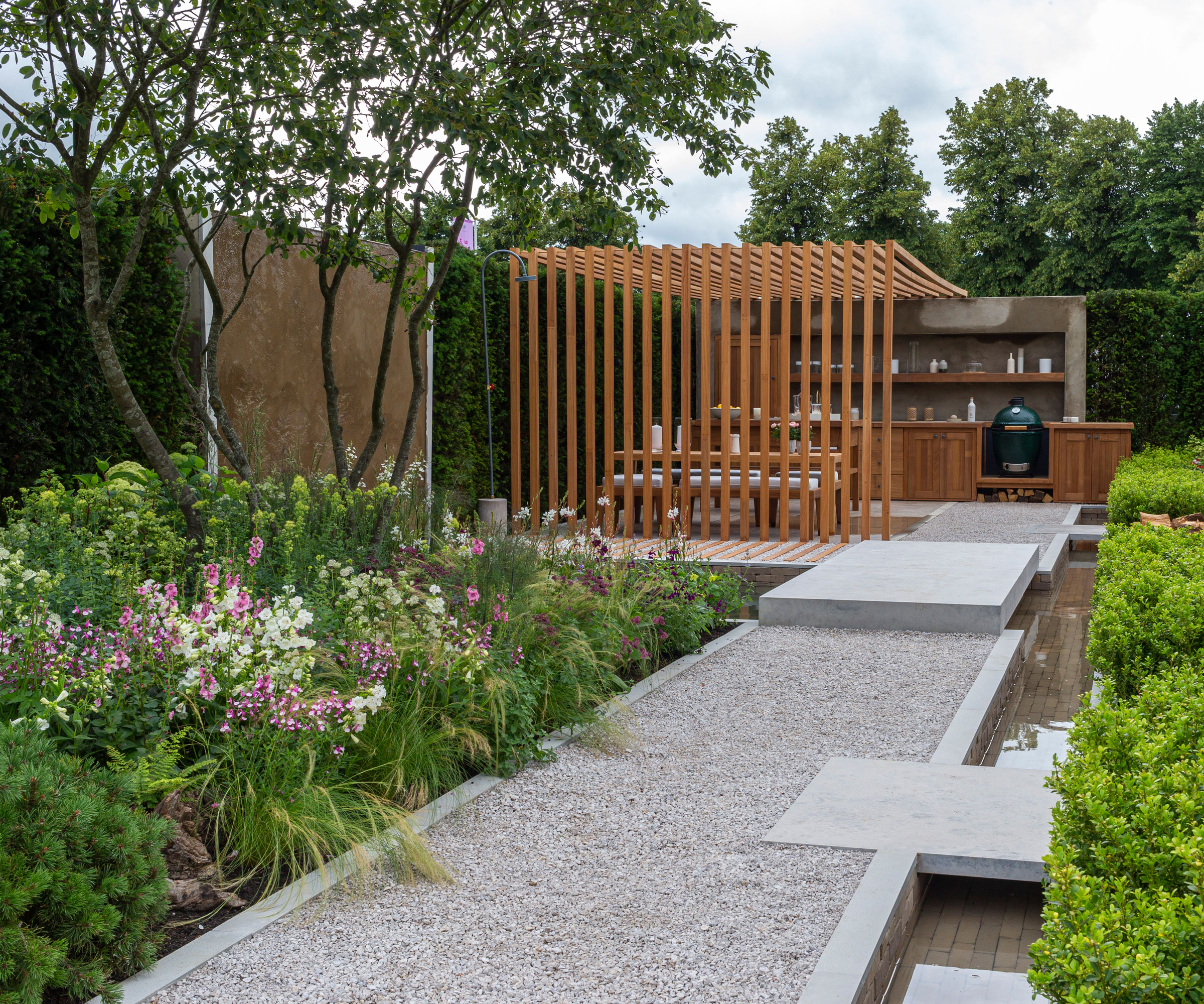 outdoor dining area and outdoor kitchen with pergola made of hardwood iroko, planting beds with white and pink flowers, clipped evergreens, water feature and gravel pathway
