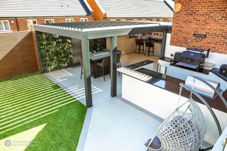 a green fixed pergola with a dining set and bar underneath it, and a huge outdoor kitchen next to it complete with BBQ, and black worktops and white cabinets