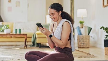 Woman in sportswear looks at smartphone, smiling. She is sitting cross-legged on the floor in a domestic setting, a towel draped over one shoulder