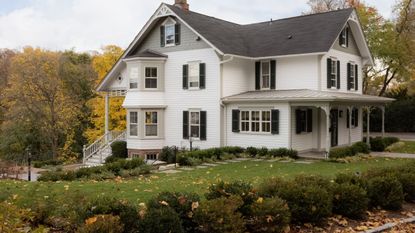 a large white historic home with sloped grey roof, window shutters, historic porch and stairs to front door surrounded by trees and lawns with fall leaves