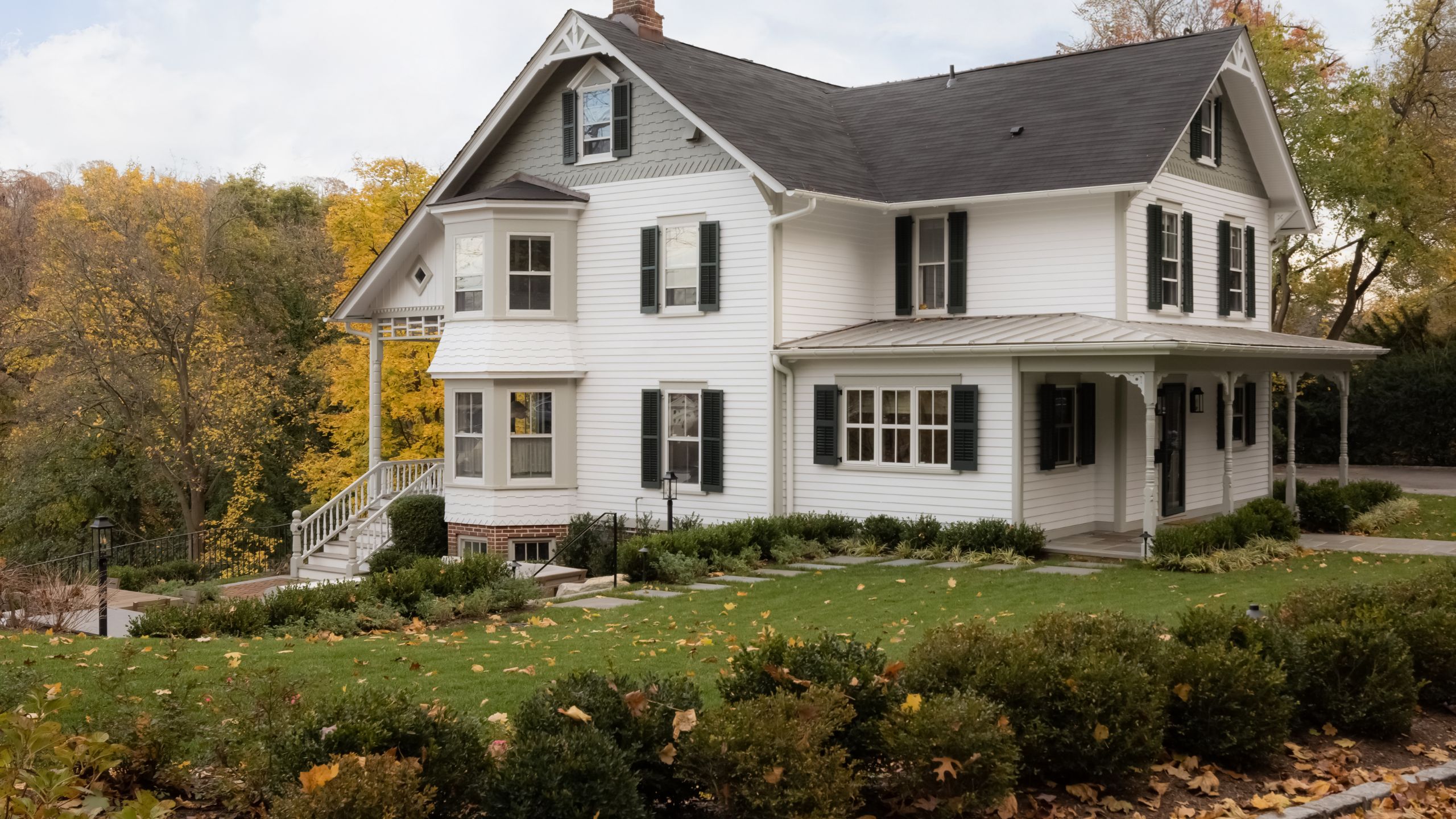 a large white historic home with sloped grey roof, window shutters, historic porch and stairs to front door surrounded by trees and lawns with fall leaves
