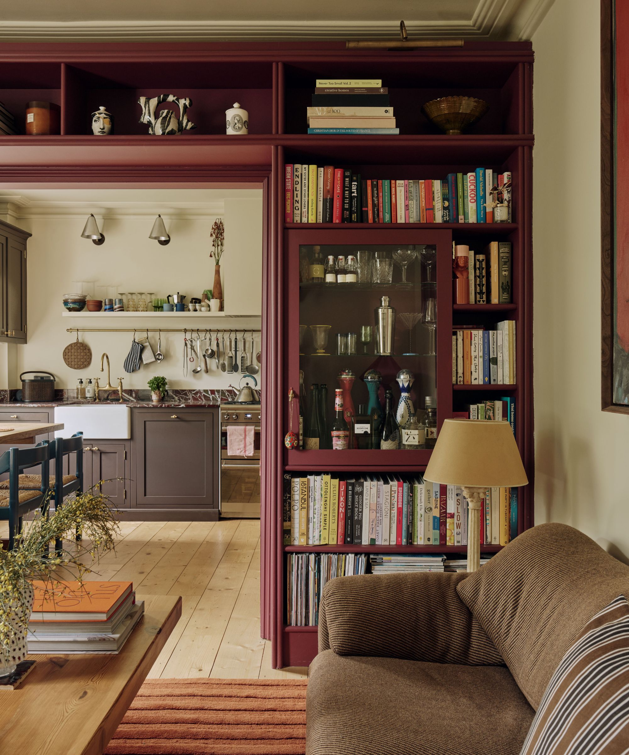 A neutral kitchen features in the background of a Living room with a burgundy built-in bookcase decorated with books and a bar area, wooden floor, striped orange rug, brown sofa and a wooden coffee table with a stack of books