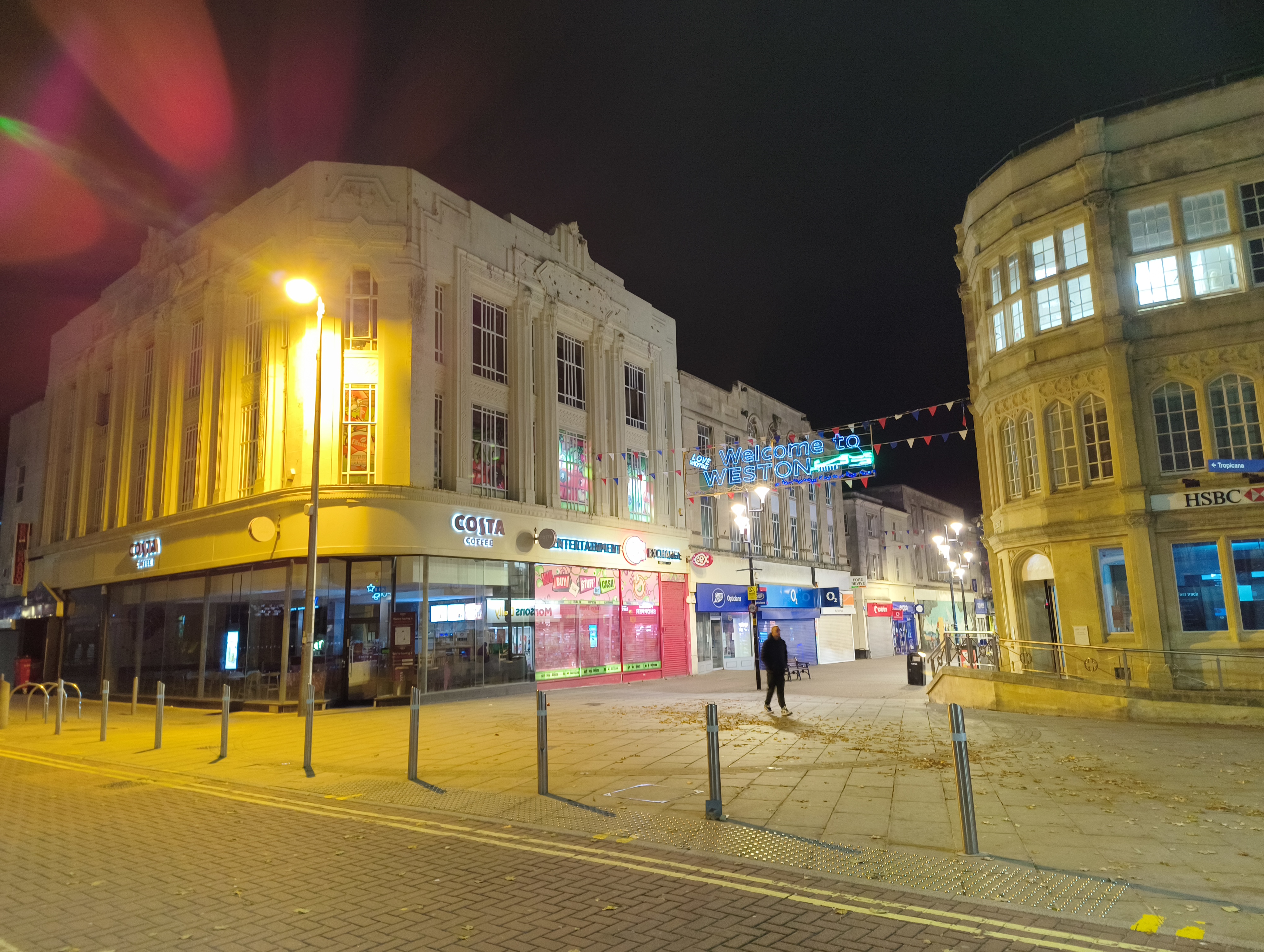 A dimly lit, nighttime scene of a town square featuring a prominent white building with a Costa Coffee on the ground floor and an older, round building with the HSBC logo on the right.