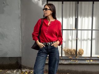 British style influencer Jessica Skye poses in front of a building wearing black oval sunglasses, a red henley sweater, a Khaite studded belt, and dark-wash barrel jeans.