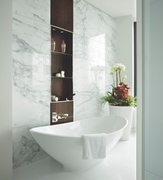 An example of spa bathroom ideas showing a marble bathroom with a walnut inset shelving unit and a white bath