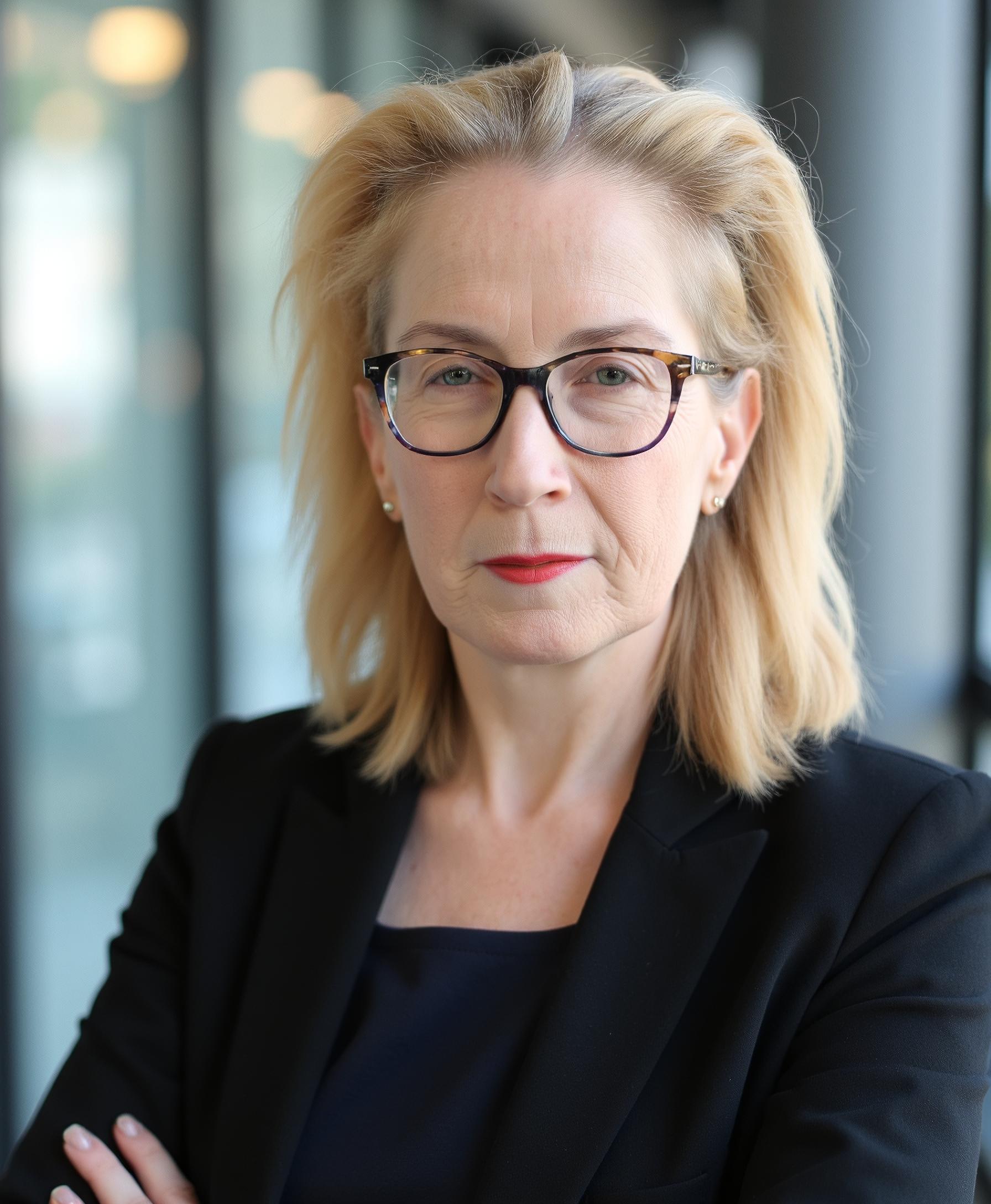a headshot of a woman with shoulder-length blonde hair wearing glasses with red lipstick staring at the camera with her arms folded and wearing a black suit