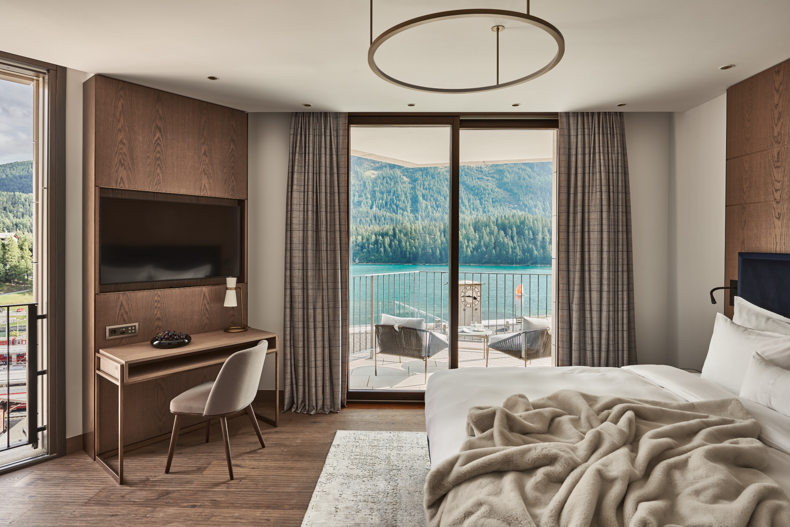 A hotel bedroom furnished in neutral tones with floor to ceiling windows with a view onto a turquoise alpine lake