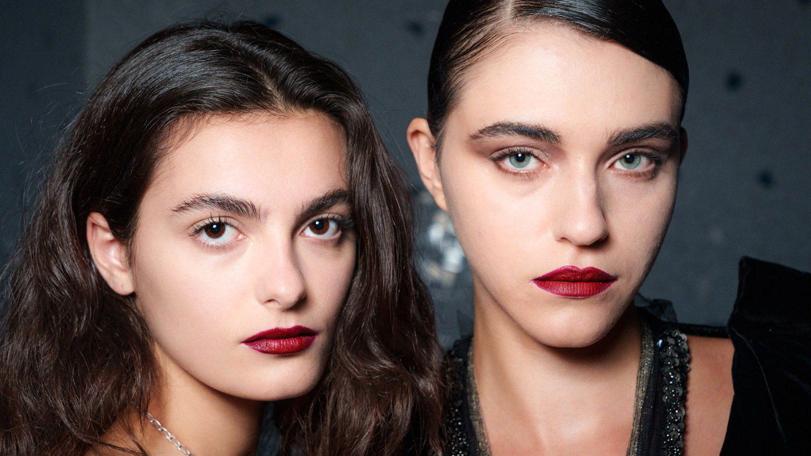 Two runway models face the camera in a close image, the models wear black clothing and their makeup consists of a claret red lip and soft brown eyeshadow with strong brows and full lashes.