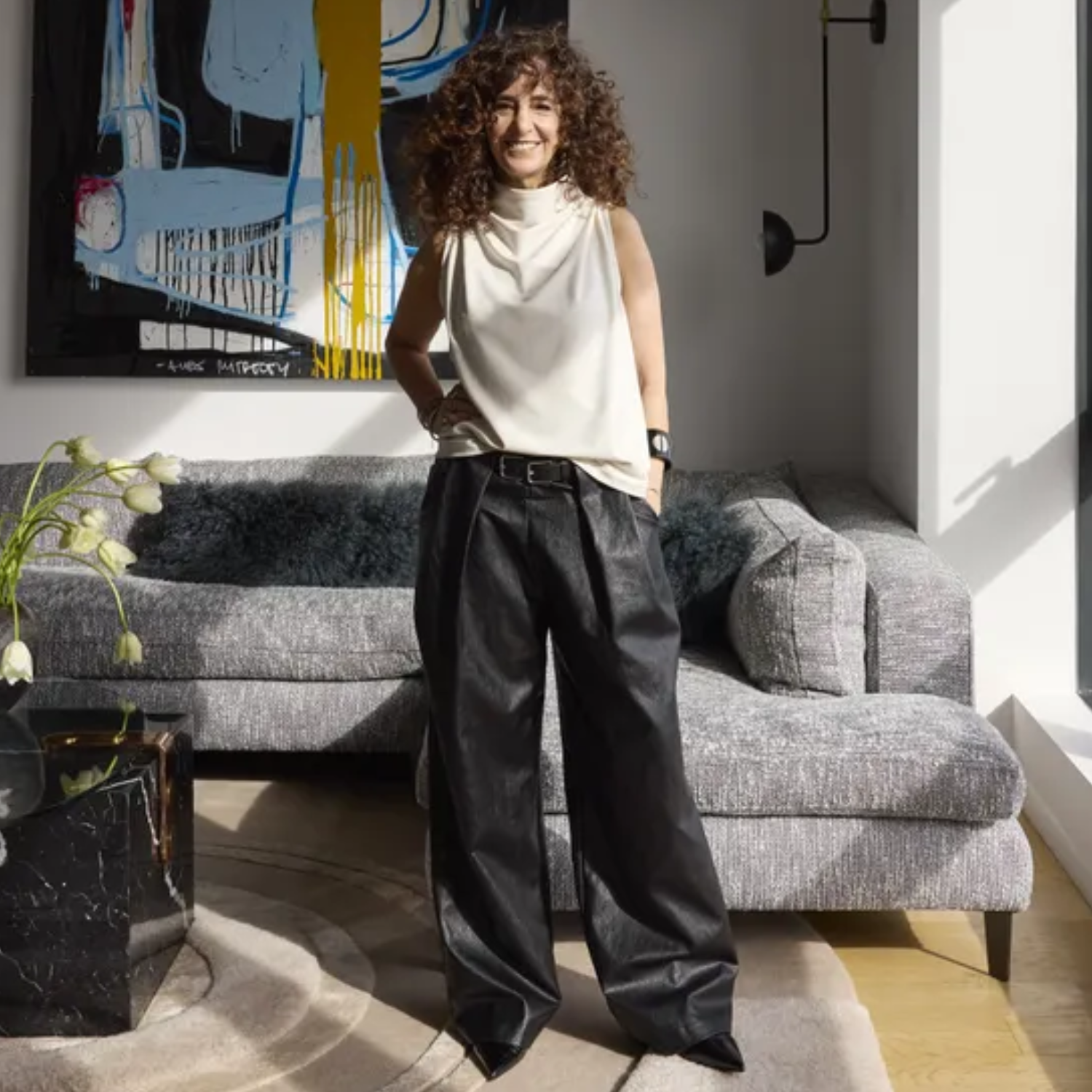 A woman is posing in a white cowl neck top and baggy black leather jeans in a white living room with a large art piece hanging in the back above a gray couch and a stone table with white tulips