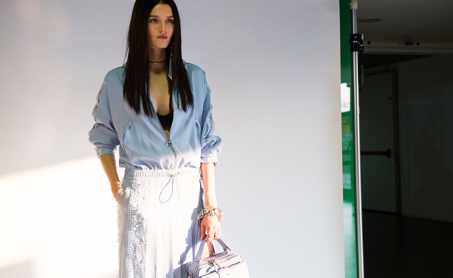 Model with sky blue jacket, white skirt and holding shrunken down handbag.