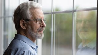 an older man with grey hair, a beard and glasses looking out a window