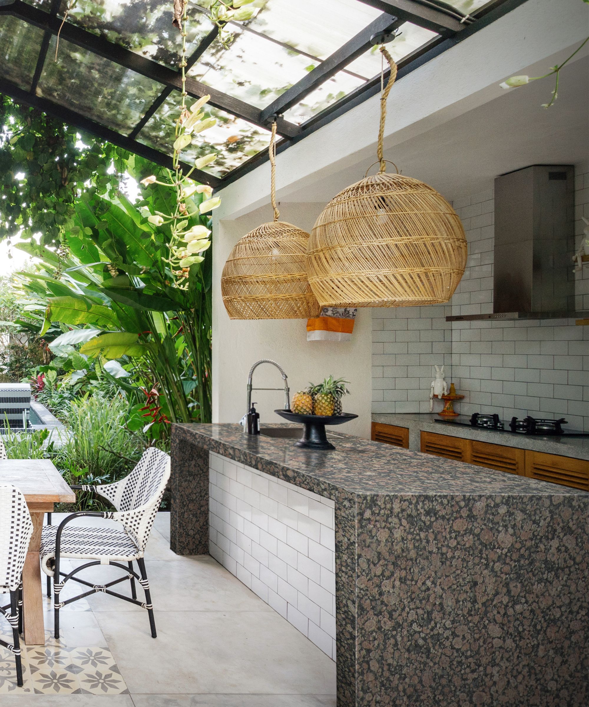A tropical outdoor kitchen under a black steel and glass roof, with a granite-topped island, large rattan globe pendants, white metro tiles, timber cabinetry and lush jungle planting beyond.
