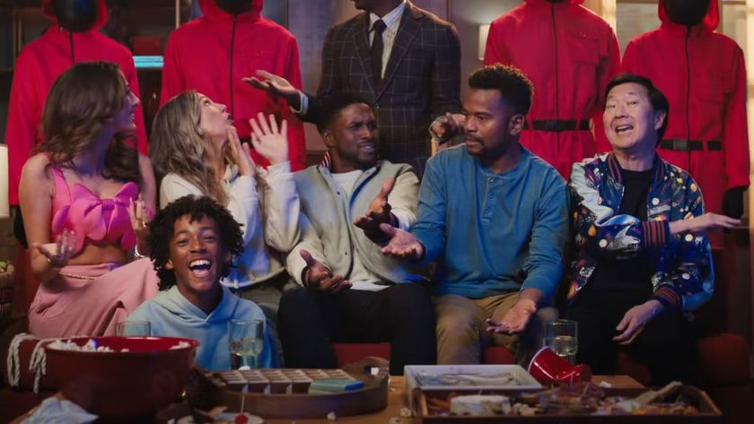 A group of people animatedly sit on a couch with Squid Game guards behind them