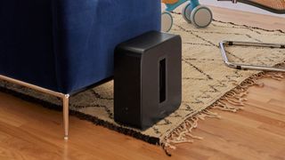 A black Sonos Sub 4 on a rug next to the end of a sofa in a lounge.