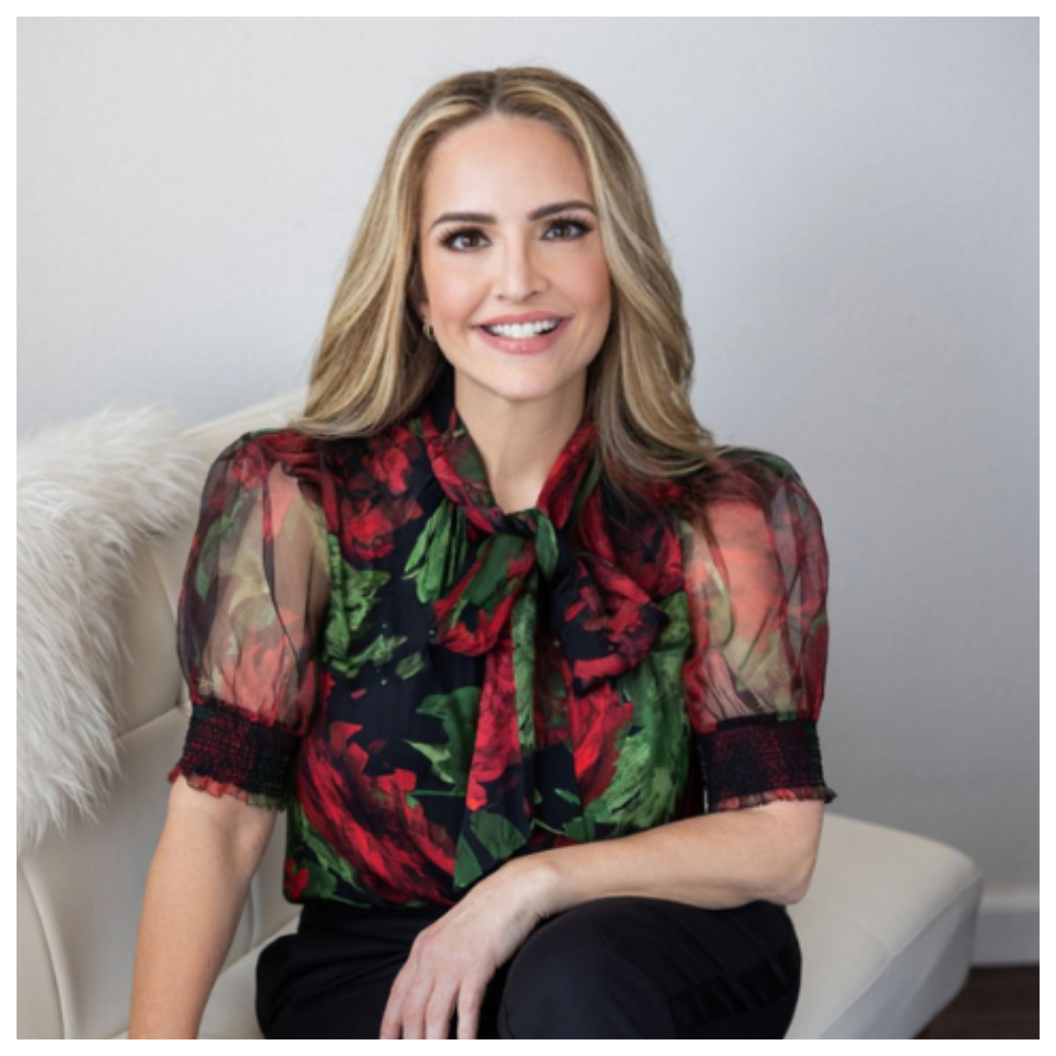 A headshot of a woman in a floral blouse with sheer puffed sleeves and black formal pants sitting cross-legged on a white sofa with a fur throw