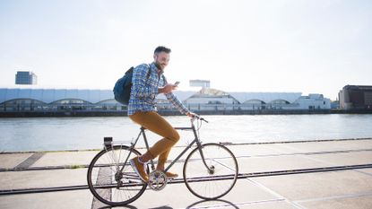 A man in casual streetware rides a flatbar bike while looking at a mobile phone in his right hand. He is smiling at the phone.