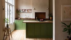 Green kitchen with bright window to the left and island in center of image. Wooden counters and accents, lower cabinets only with chimney breast over range cooked in background