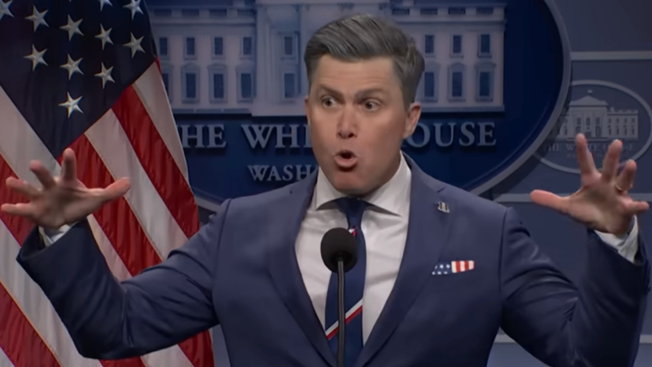 Colin Jost dressed in a suit as Pete Hegseth gives a press conference.