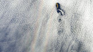 A satellite photo showing a faint double band of rainbow next to an island with cloud swirls forming off its coastline