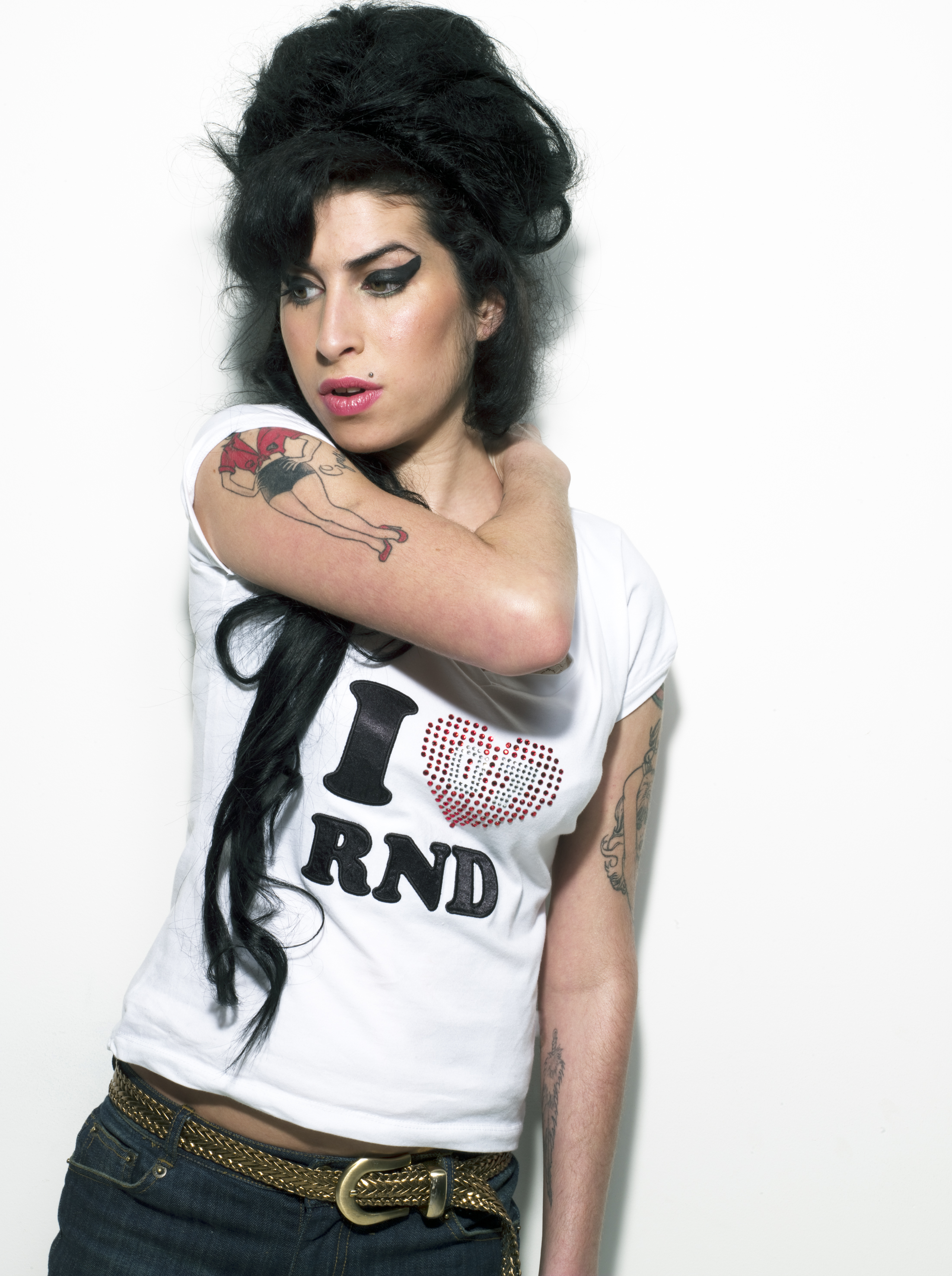 Amy Winehouse stands against a white background in a white crop top and jeans, her signature beehive and heavy eye makeup in place, arms folded and gaze directed away from camera.