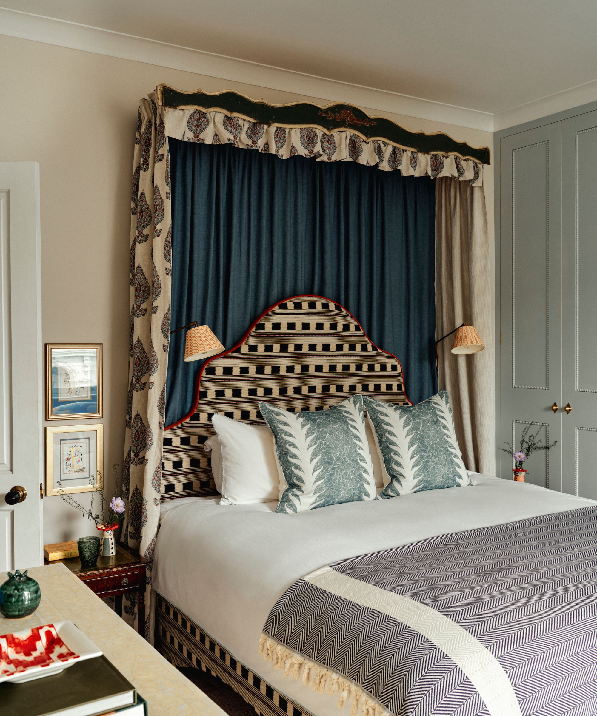 A well-appointed bedroom with neutral walls and light blue paneled wardrobes. The bed features a unique arched headboard with horizontal black and white stripes and red piping. Above the bed is a grand canopy consisting of a dark, ornate wooden valance and blue drapes. Two small, pleated sconce lamps are mounted on either side of the headboard. The bed is dressed in crisp white linens, teal patterned pillows, and a gray herringbone throw.