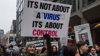 photo shows a man outdoors at a protest in London holding a sign that reads "It's not about a 'virus,' it's about control."