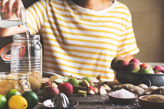 A close-up of a women pouring water into a jar, surrounded by fruit and vegetables.