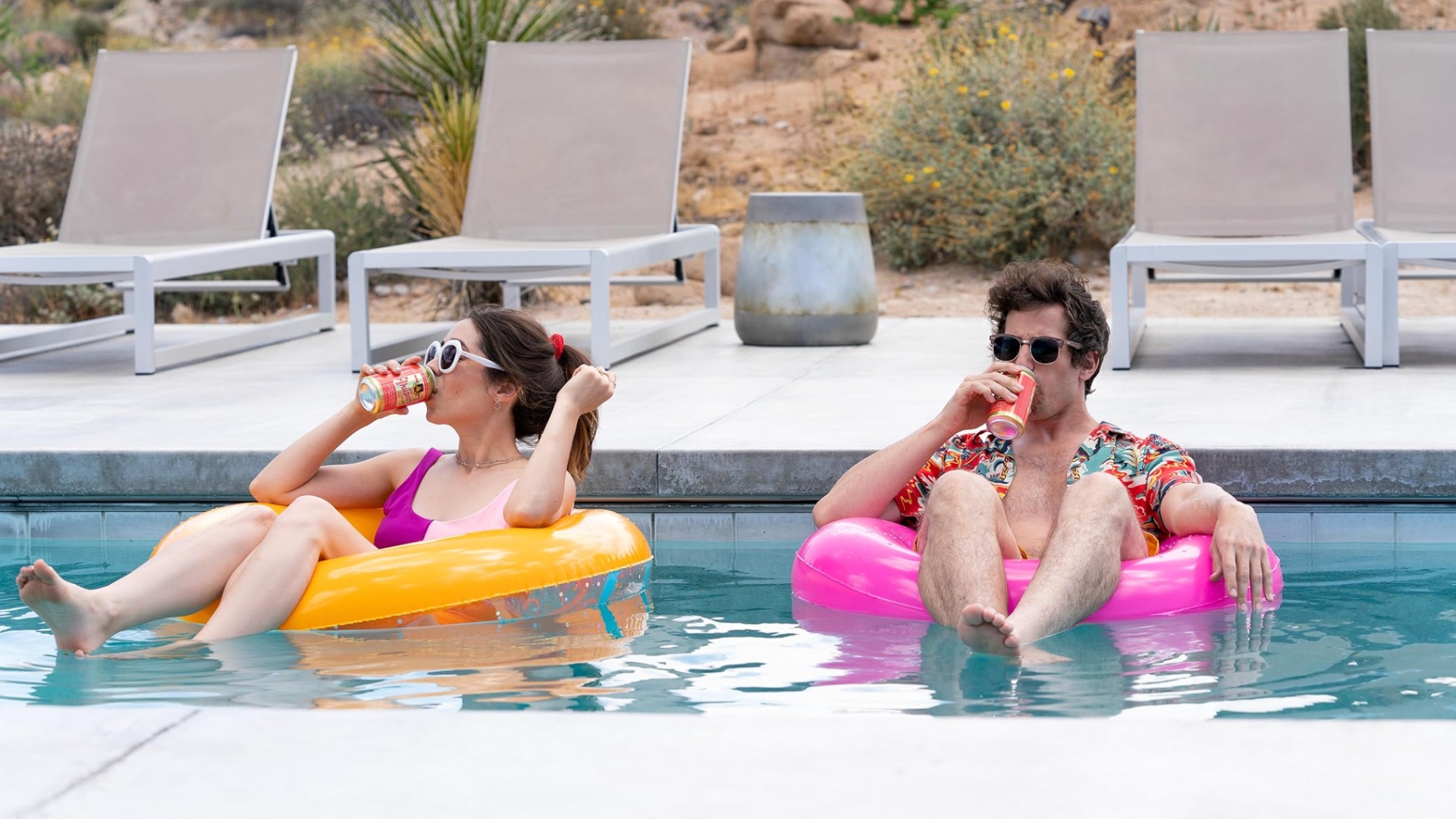 A dark haired man and woman, both sat in pool rings in a swimming pool, drinking cans of a beverage.