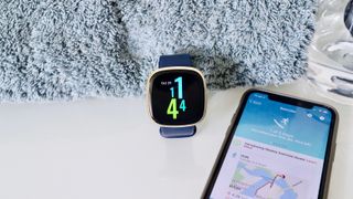 En Fitbit Versa 3 ligger bredvid en mobil med Fitbit-appen uppe, på ett ljus skrivbord och en grå filt.