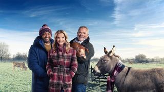 JB Gill, Helen Skelton and Jules Hudson stand against a backdrop of a field with Jules holding a piglet and a donkey stands next to them in Winter on the Farm.