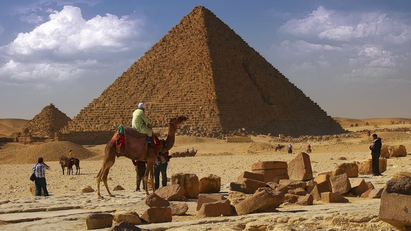A few people and a camel are before a large Egyptian pyramid on a blue sky day.