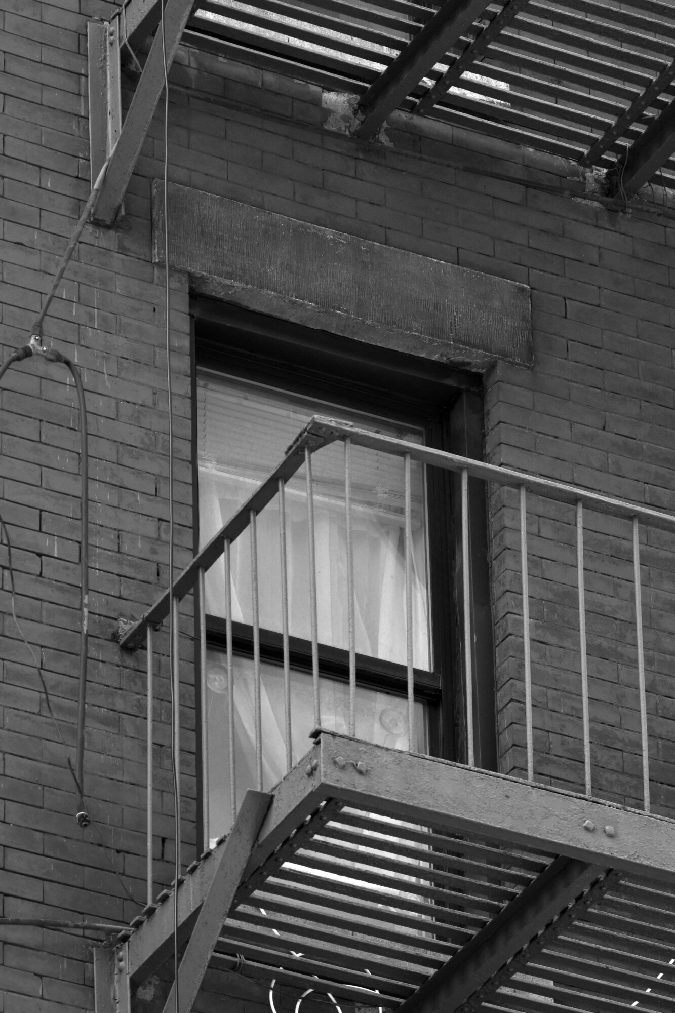 A brick apartment window behind a fire escape, with net curtains drawn and nobody in sight.