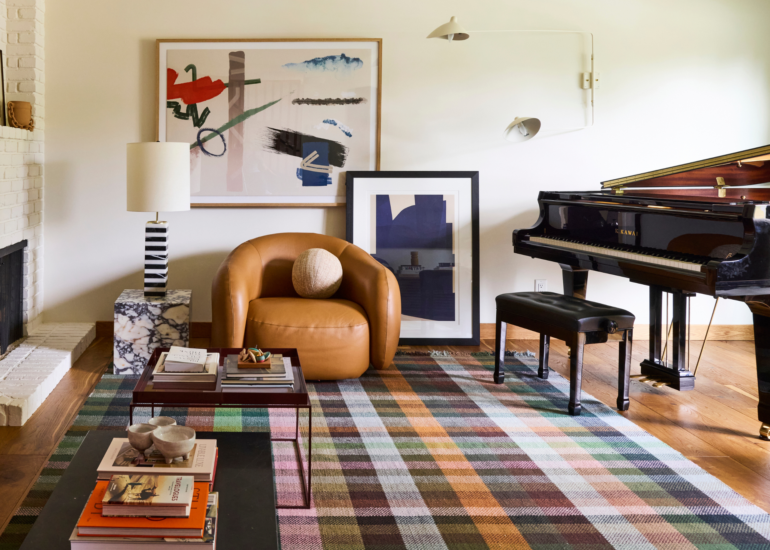 A checked multi-color rug with a leather couch, two coffee tables, a grand piano, a leaning painting, and a plinth with a striped table lamp