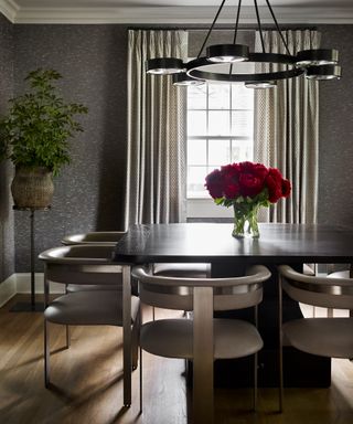 Dining room with wood floor and gray wallpaper, upholstered dining chairs, dark wood table, vase of red roses, plant on stand, pendant light and window dressed with curtains