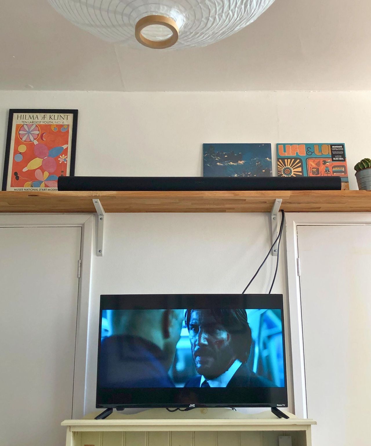 Sonos Arc Ultra soundbar in Dan Fauzi's home, on wooden shelf with cactus plant, above TV showing John Wick: Chapter 2. A white lampshade is pictured in the top left corner, above two prints on the wall.